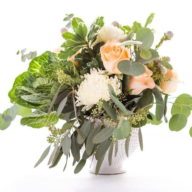 A fresh floral arrangement featuring peach roses, white chrysanthemums, and green eucalyptus leaves in a textured white ceramic pot, set against a solid white background.