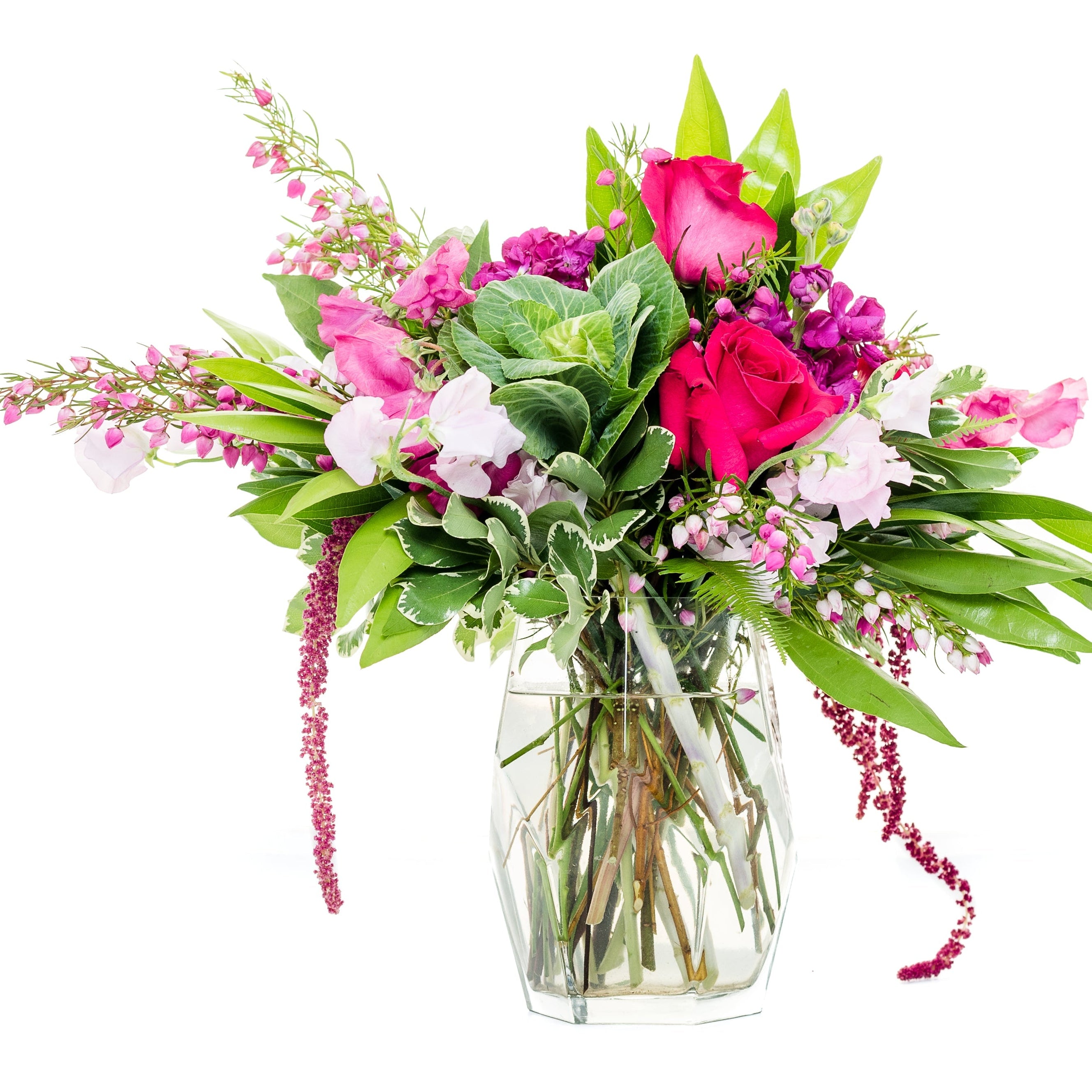 A vibrant mixed floral bouquet in a clear glass vase, featuring bright pink roses, magenta blossoms, white flowers, and lush green foliage, isolated on a solid white background.