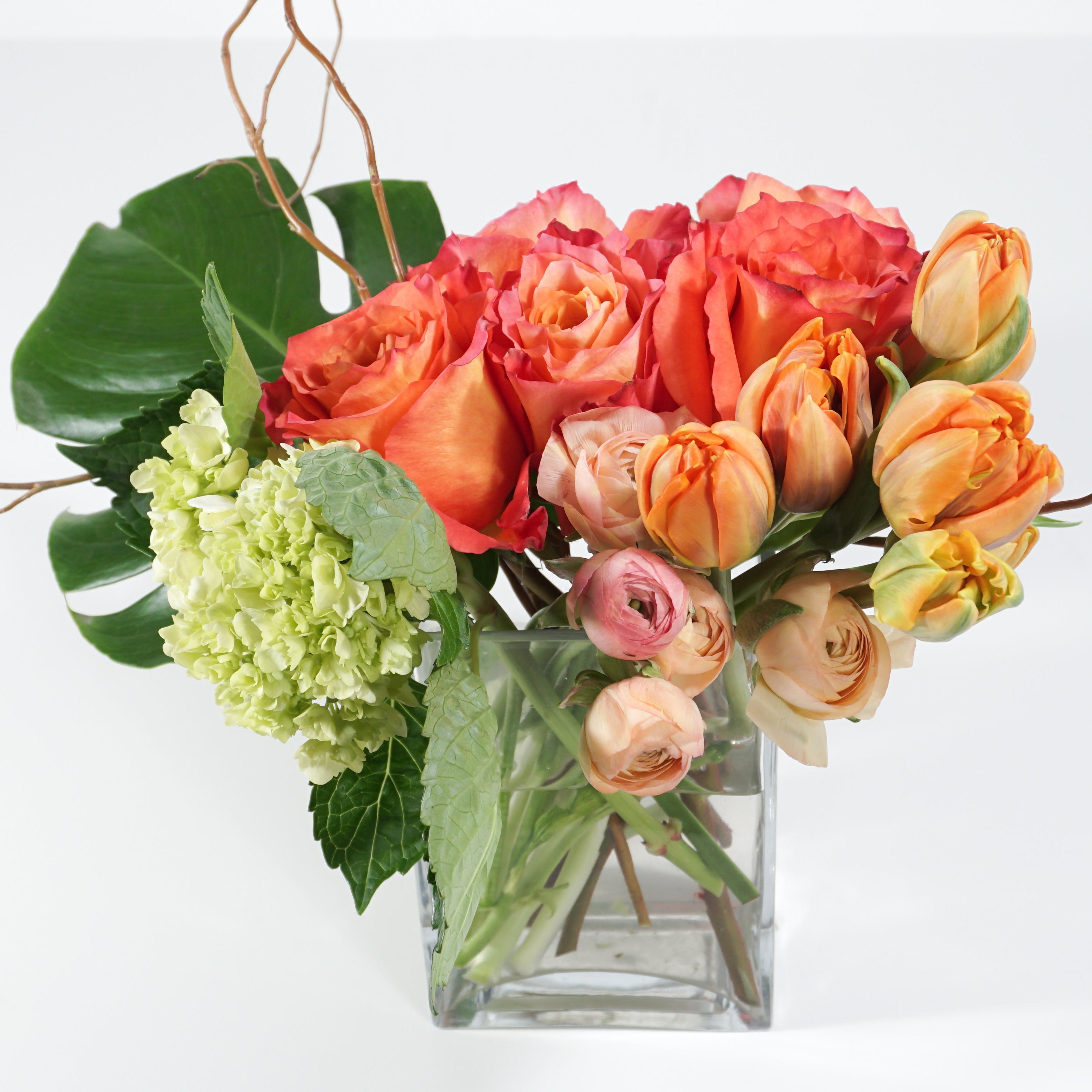 A floral arrangement featuring orange and coral roses, peach tulips, pale pink ranunculus, and light green hydrangea blooms in a clear square glass vase on a white background.